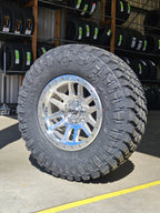 A large off-road tire mounted on a multi-spoke silver chrome wheel, photographed in a warehouse setting with other tires on shelves in the background.